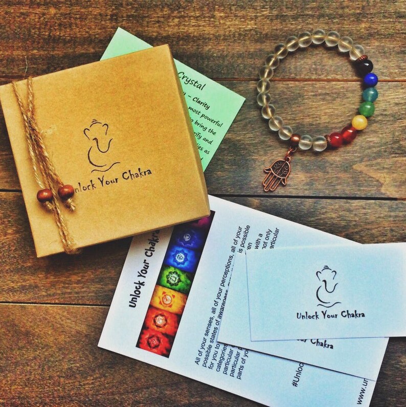 May include: A clear crystal bracelet with a hamsa charm and seven chakra colored beads. The bracelet is packaged in a brown box with the text "Unlock Your Chakra" on it. The box is sitting on a wooden surface.