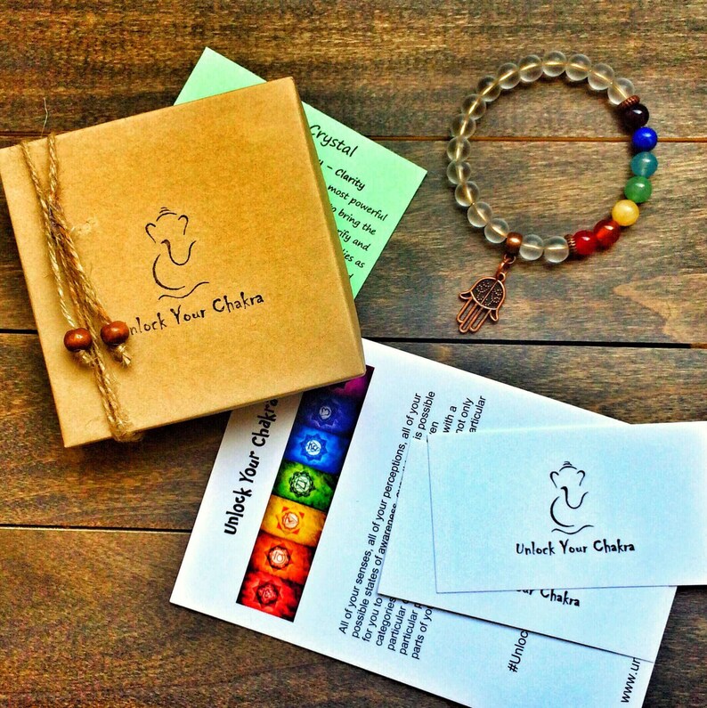 A clear crystal bracelet with a copper Hamsa charm and seven chakra colored beads. The bracelet is on a wooden surface with a small brown box and two cards that say "Unlock Your Chakra".