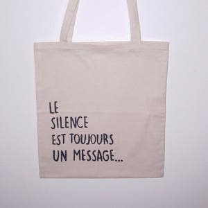 Op de afbeelding: Een beige draagtas met lange handvatten. De tas heeft de Franse tekst "LE SILENCE EST TOUJOURS UN MESSAGE..." in zwarte letters. De tas is gemaakt van een lichtgekleurde stof en hangt tegen een witte achtergrond.