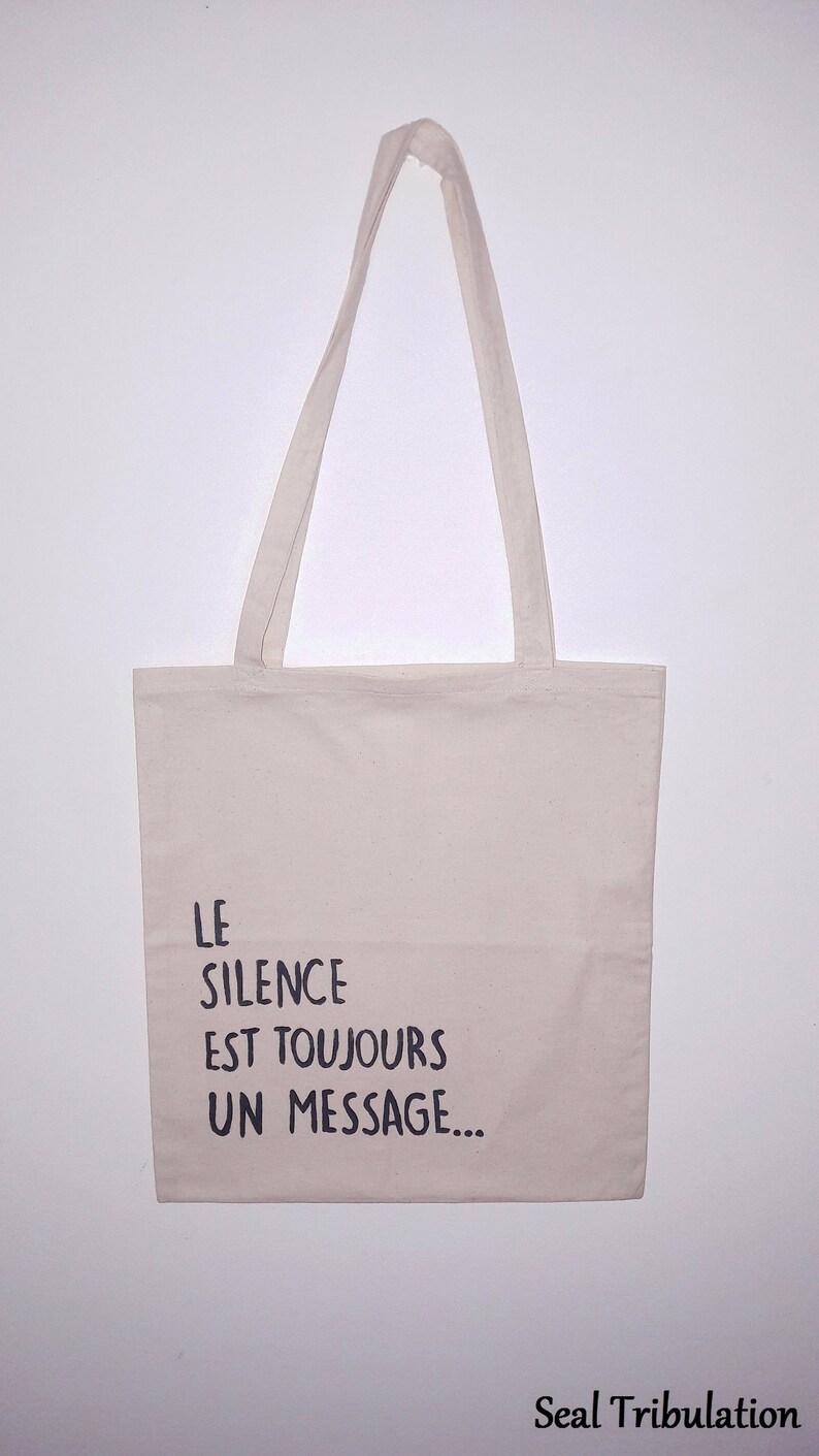 Op de afbeelding: Een beige draagtas met lange handvatten. De tas heeft de Franse tekst "LE SILENCE EST TOUJOURS UN MESSAGE..." in zwarte letters. De tas is gemaakt van een lichtgekleurde stof en hangt tegen een witte achtergrond.