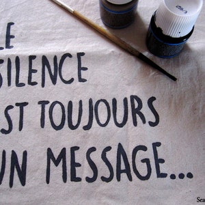 Op de afbeelding: Een beige canvas met de Franse tekst "LE SILENCE EST TOUJOURS UN MESSAGE..." in zwarte letters. Een penseel en twee flessen zwarte verf liggen op het canvas. De tekst "Seal Tribulation" staat in de rechterbenedenhoek.