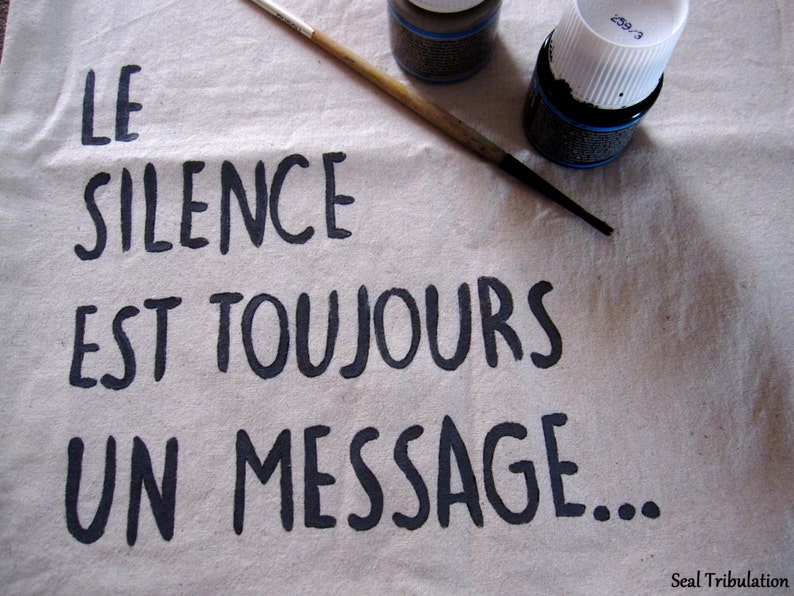 Op de afbeelding: Een beige canvas met de Franse tekst "LE SILENCE EST TOUJOURS UN MESSAGE..." in zwarte letters. Een penseel en twee flessen zwarte verf liggen op het canvas. De tekst "Seal Tribulation" staat in de rechterbenedenhoek.