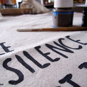 Op de afbeelding: Close-up van een canvas met de woorden "LE SILENCE" in zwarte verf. Een penseel en twee kleine verfpotjes staan op de achtergrond. Het canvas is gebroken wit.