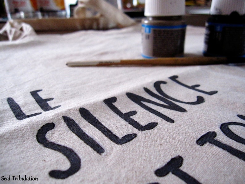 Op de afbeelding: Close-up van een canvas met de woorden "LE SILENCE" in zwarte verf. Een penseel en twee kleine verfpotjes staan op de achtergrond. Het canvas is gebroken wit.
