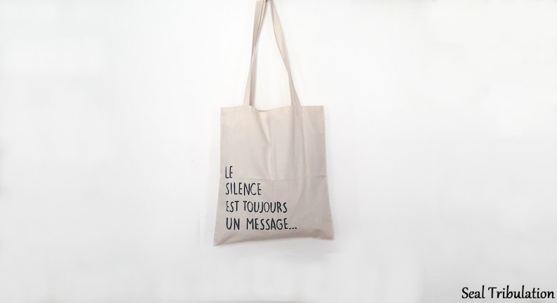 Op de afbeelding: Beige draagtas met lange hengsels, hangend tegen een witte achtergrond. De tas heeft de Franse tekst "LE SILENCE EST TOUJOURS UN MESSAGE..." in zwarte letters, wat "Stilte is altijd een boodschap" betekent.