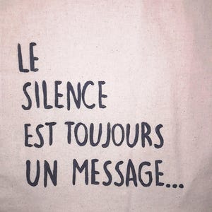 Op de afbeelding: Een beige canvas draagtas met de Franse tekst "LE SILENCE EST TOUJOURS UN MESSAGE..." in donkergrijze letters. De tas heeft een eenvoudig ontwerp.