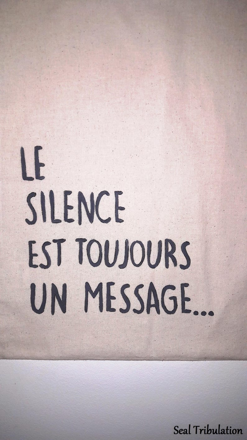 Op de afbeelding: Een beige canvas draagtas met de Franse tekst "LE SILENCE EST TOUJOURS UN MESSAGE..." in donkergrijze letters. De tas heeft een eenvoudig ontwerp.