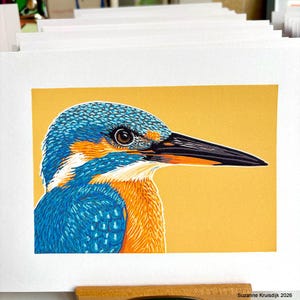 Op de afbeelding: Een rechthoekige kunstdruk met een illustratie van een ijsvogel tegen een effen gele achtergrond. De vogel heeft felblauwe veren, een oranje borst en een lange zwarte snavel. De tekst Suzanne Kruisdijk 2026 staat in de rechterbenedenhoek.