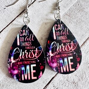 May include: A pair of teardrop-shaped earrings with a black background and a galaxy-like design. The earrings feature the text "I can do all things through Christ who strengthens me".
