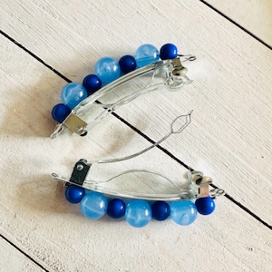 May include: Two silver-toned hair barrettes, each adorned with a row of blue beads. The beads alternate between solid and translucent shades of blue. The barrettes are set against a white wooden surface.