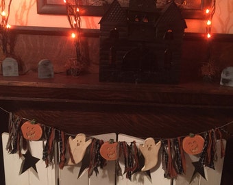 May include: Halloween-themed mantel decoration featuring a black haunted house, string lights, and a garland with pumpkins, ghosts, and stars. The garland is draped over white shutters, creating a festive display.