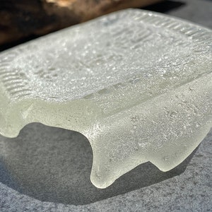 May include: A clear, frosted glass piece with a slightly irregular shape. The glass has a textured surface and appears to be sea glass.