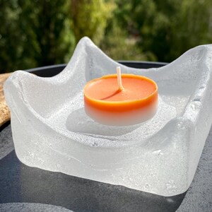 May include: A clear, textured glass dish with an irregular, wavy rim holding a small, round tea light candle. The candle features a vibrant orange top half and a white bottom half, with a single white wick centered in the middle of the wax.