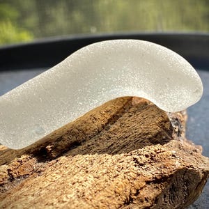 May include: A piece of sea glass, translucent and curved, rests on a piece of weathered driftwood. The sea glass is a soft, frosted white, and the wood is a rich brown with a textured surface. The background is blurred, suggesting a natural setting.