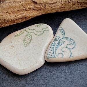 May include: Two off-white, smooth stones with decorative green and teal floral patterns. One stone is oval-shaped, the other is triangular. The stones are placed on a dark gray surface, with a piece of driftwood in the background.
