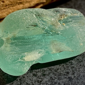 May include: A smooth, light green sea glass piece with a slightly cloudy appearance. The glass has a unique, organic shape and is likely a naturally tumbled piece of sea glass.