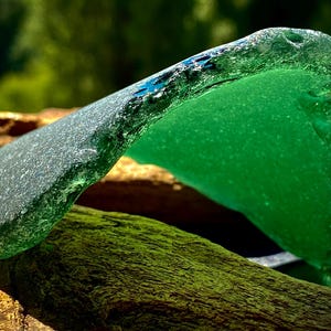 May include: A piece of sea glass with a green hue and a rough, textured surface. The glass is shaped like a wave and has a translucent quality.