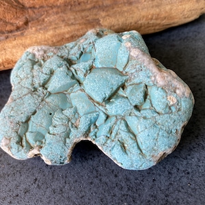 Puede incluir: Una piedra de color turquesa, de forma irregular, con una superficie texturizada y grietas visibles. La piedra tiene una mezcla de tonos azules claros y oscuros, con algunas áreas que parecen más claras. Un trozo de madera flotante marrón está en el fondo.