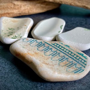 Puede incluir: Cuatro piezas de cerámica marina en tonos blanco, beige y verde claro. Una pieza presenta un diseño turquesa. La cerámica es lisa y redondeada, con un aspecto desgastado. Las piezas están dispuestas sobre una superficie oscura.