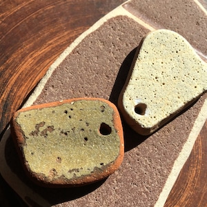 以下が含まれることがあります： 穴の開いた、形が不規則な2つのテクスチャードセラミックペンダント。1つはライトベージュ、もう1つは緑と茶色の表面です。茶色とベージュのセラミックベースの上にあり、暗い茶色のテクスチャード背景があります。