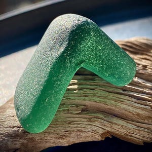 May include: A green sea glass piece shaped like a boomerang, resting on a piece of driftwood.