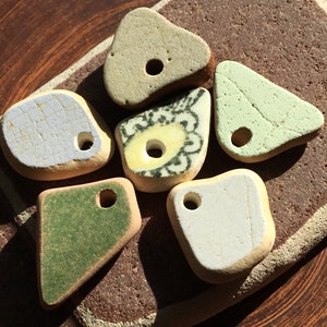 Puede incluir: Colección de seis piezas de cerámica marina de diversas formas y colores. Incluye un triángulo verde claro, una piedra beige y una pieza con diseño floral. Cada pieza tiene un agujero perforado.