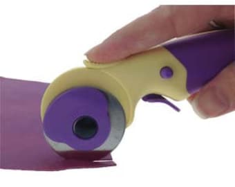 May include: Purple and yellow rotary cutter with a safety lock and a blade guard. The cutter is being used to cut a piece of purple fabric.