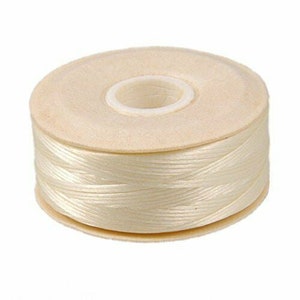 Pode incluir: Uma bobina de linha creme. A linha é enrolada firmemente em torno de um núcleo de cartão. A bobina é redonda e está sobre um fundo branco. Adequado para projetos de costura e artesanato.