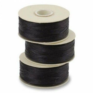 May include: Three stacked spools of black thread. Each spool has a beige cardboard core and is tightly wound with the dark thread. The spools are arranged in a vertical tower, showcasing the thread's texture and color.
