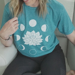 May include: Teal t-shirt with a white floral design and moon phase graphics. The shirt is a crew neck and has short sleeves. The design is centered on the front of the shirt.