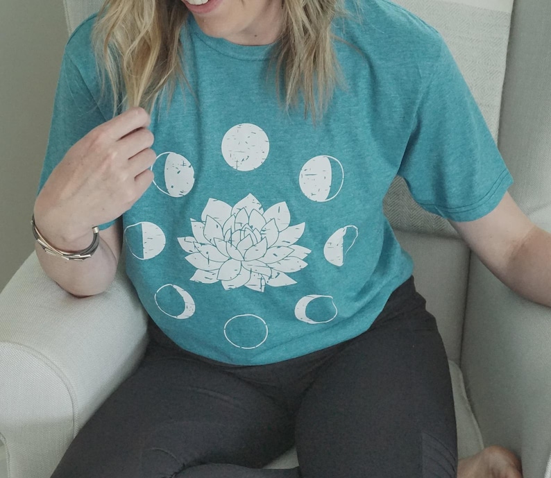 May include: Teal t-shirt with a white floral design and moon phase graphics. The shirt is a crew neck and has short sleeves. The design is centered on the front of the shirt.
