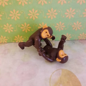 May include: Two brown plastic toy monkeys with yellow faces and ears. The monkeys are in a playful pose, one on top of the other.