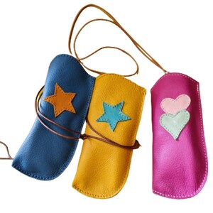 Può includere: Tre custodie per occhiali in pelle con un cinturino. Le custodie sono blu, gialla e rosa. La custodia blu ha una stella gialla, la custodia gialla ha una stella blu e la custodia rosa ha due cuori rosa.
