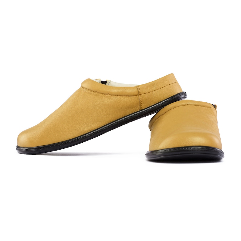 May include: A pair of yellow leather slippers with black soles. The slippers are laid out on a white background.