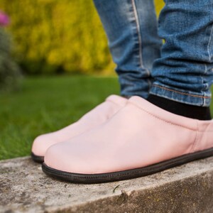 May include: A pair of pink leather slip-on shoes with black soles. The shoes are worn by a person wearing blue jeans.