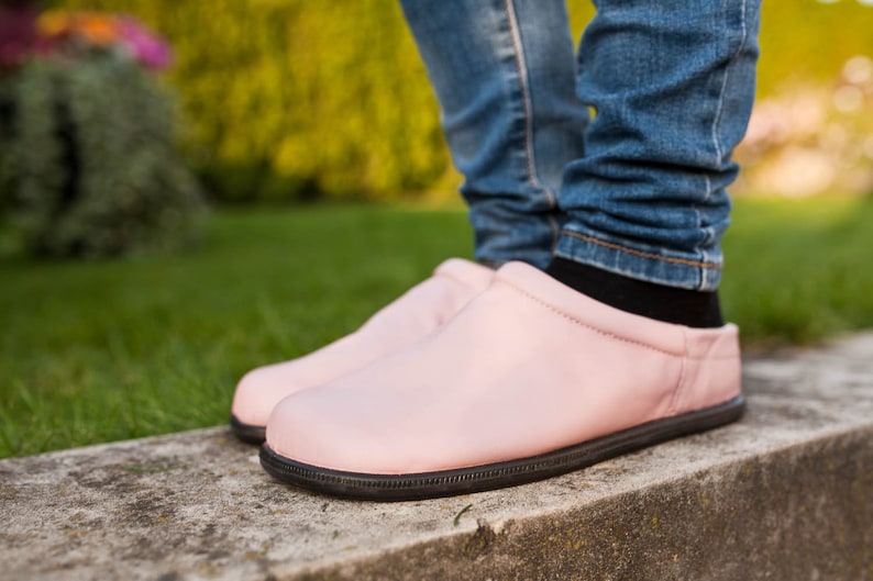 May include: A pair of pink leather slip-on shoes with black soles. The shoes are worn by a person wearing blue jeans.