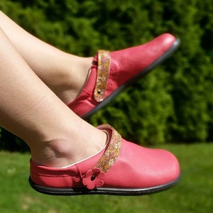 May include: A pair of pink leather slip-on shoes with floral patterned straps. The shoes have a black rubber sole.