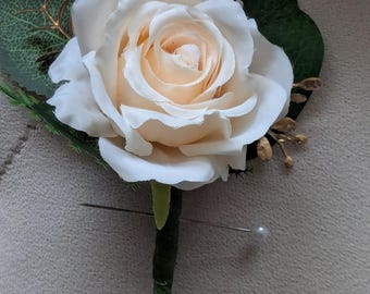 May include: A single white rose with gold accents and green leaves, attached to a pin with a pearl head.