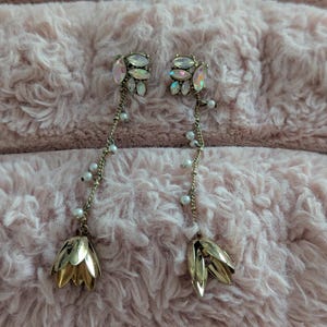 May include: A pair of long drop earrings featuring a cluster of iridescent marquise-cut crystals at the top, connected by a delicate gold-tone chain adorned with small white beads, ending in a gold-tone metal flower-shaped pendant at the base.