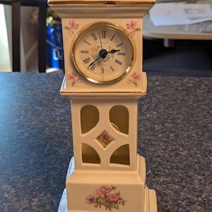 May include: A white ceramic grandfather clock with gold trim, featuring a round clock face with Roman numerals, pink floral accents, and a cutout design on the lower section. The clock face includes the text Quartz and a floral graphic.