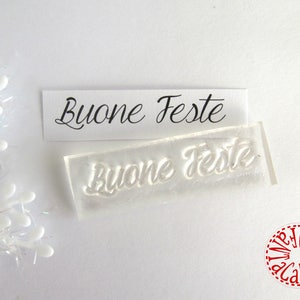 Może przedstawiać: Przezroczysty stempel akrylowy i wydrukowana karta ze słowami "Buone Feste" w eleganckim czarnym skrypcie. Stempel ma kształt prostokąta z tekstem w wypukłym reliefie. Element dekoracyjny znajduje się w tle.