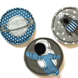 Könnte beinhalten: Drei runde Stoffpatches mit Weltraummotiven. Ein Patch zeigt einen blauen Planeten mit einem silbernen Ring, ein anderer Patch zeigt eine graue Rakete mit blau-weißen Streifen und der dritte Patch zeigt einen grauen Astronauten in einem blauen Raumanzug.