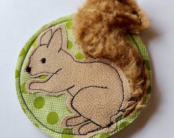 May include: A brown and tan embroidered squirrel with a fluffy brown tail on a green and white polka dot background.