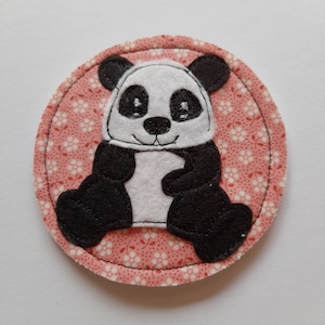 Könnte beinhalten: Ein runder, rosa Stoffuntersetzer mit einer Applikation eines schwarz-weißen Pandabären. Der Panda sitzt mit seinen Pfoten vor sich und hat eine schwarze Nase und Augen.