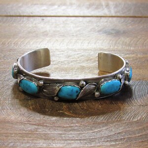 May include: A silver cuff bracelet with oval turquoise stones and leaf accents. The open-ended bracelet is displayed on a wooden surface. The turquoise stones are a vibrant blue color, and the bracelet has a vintage aesthetic.