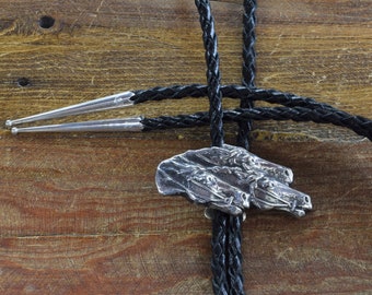 May include: A silver bolo tie featuring a detailed horse head design. The pendant is attached to a black braided leather cord with silver tips. The background is a wooden surface.