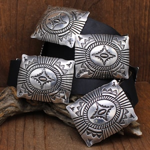 May include: Four silver belt buckles with intricate geometric designs. Each buckle features a central star-shaped motif surrounded by concentric circles and radiating lines. The buckles are arranged on a black leather belt.