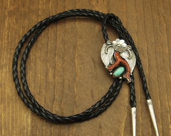 May include: A silver bolo tie with a turquoise stone and coral accents. The bolo tie features a braided black leather cord.