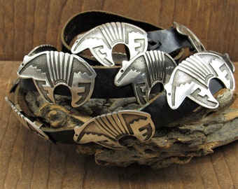 May include: A silver belt buckle with a Native American design of a bear. The buckle is on a black leather belt. The belt is on a piece of wood.