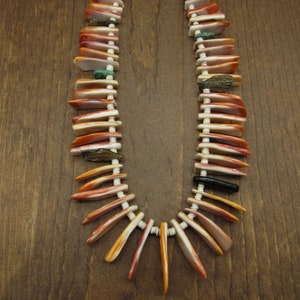 May include: A Native American style necklace made of orange, white, and brown shell pieces. The necklace is strung on a white cord and has a black bead at the bottom.